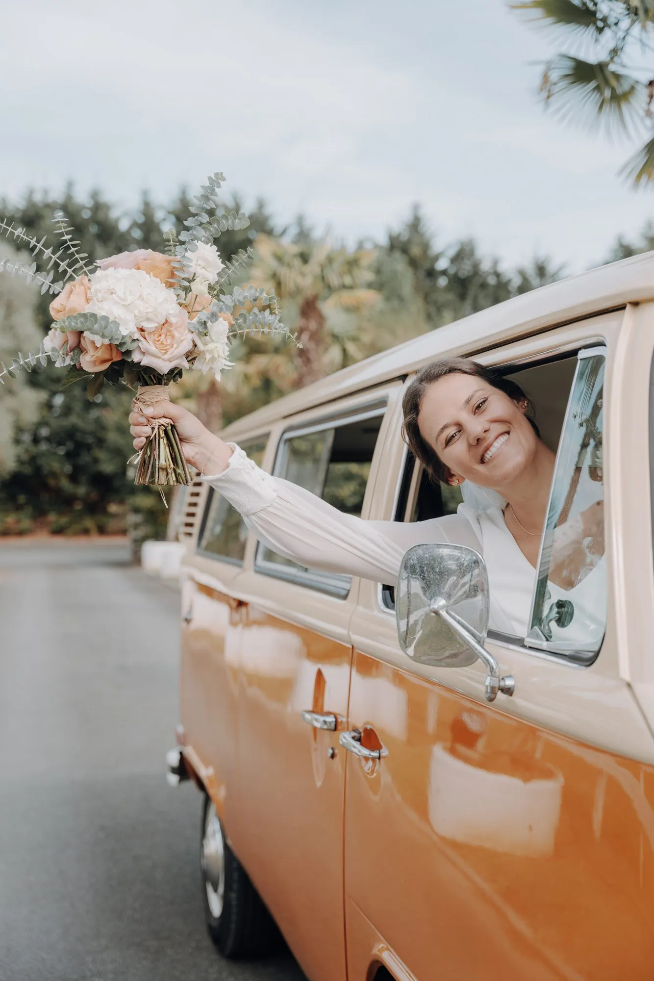 La mariée salue depuis un combi Volkswagen orange, bouquet à la main