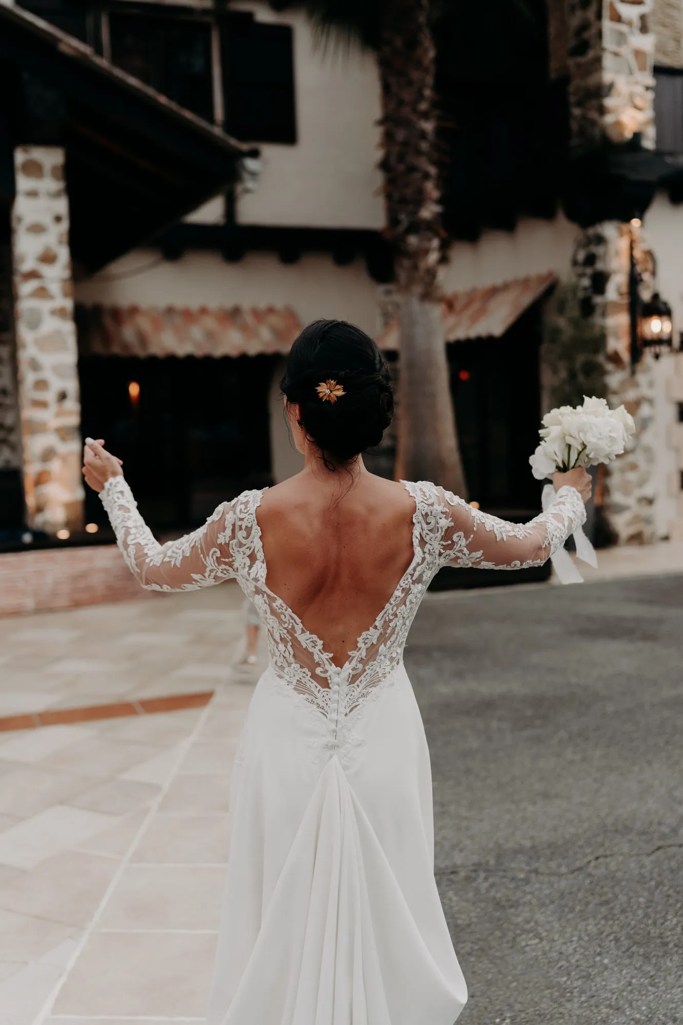 La mariée de dos, robe en dentelle, bouquet levé dans la cour du Domaine